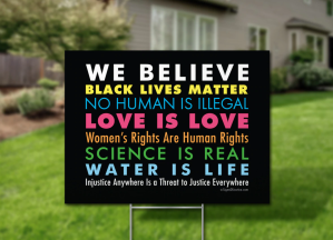 Lawn sign in front of large house reading: WE BELIEVE/ BLACK LIVES MATTER/NO HUMAN IS ILLEGAL/LOVE IS LOVE/Women's Rights Are Human Rights/SCIENCE IS REAL/WATER IS LIFE/Injustice Anywhere Is a Threat to Justice Everywhere. Copyright SignsOfJustice.com